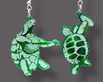 May include: A pair of green acrylic earrings featuring a turtle playing a banjo and a turtle with a shell pattern. The earrings are attached to silver hooks.