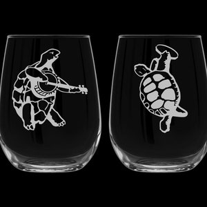 May include: Two clear glass wine glasses with etched designs of turtles. One turtle is playing a banjo and the other is upside down.