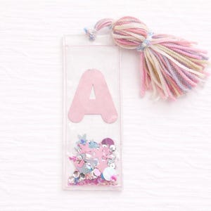 May include: A clear, rectangular bookmark with a pink letter "A" and colorful confetti. A tassel made of pink, yellow, and blue yarn is attached to the top. The bookmark is a decorative accessory.