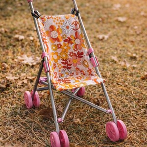 May include: A toy doll stroller featuring a metal frame with pink plastic handles and wheels. The seat fabric displays a repeating pattern of orange, yellow, and white flowers, along with peace signs, on a light background with a simple folding design.