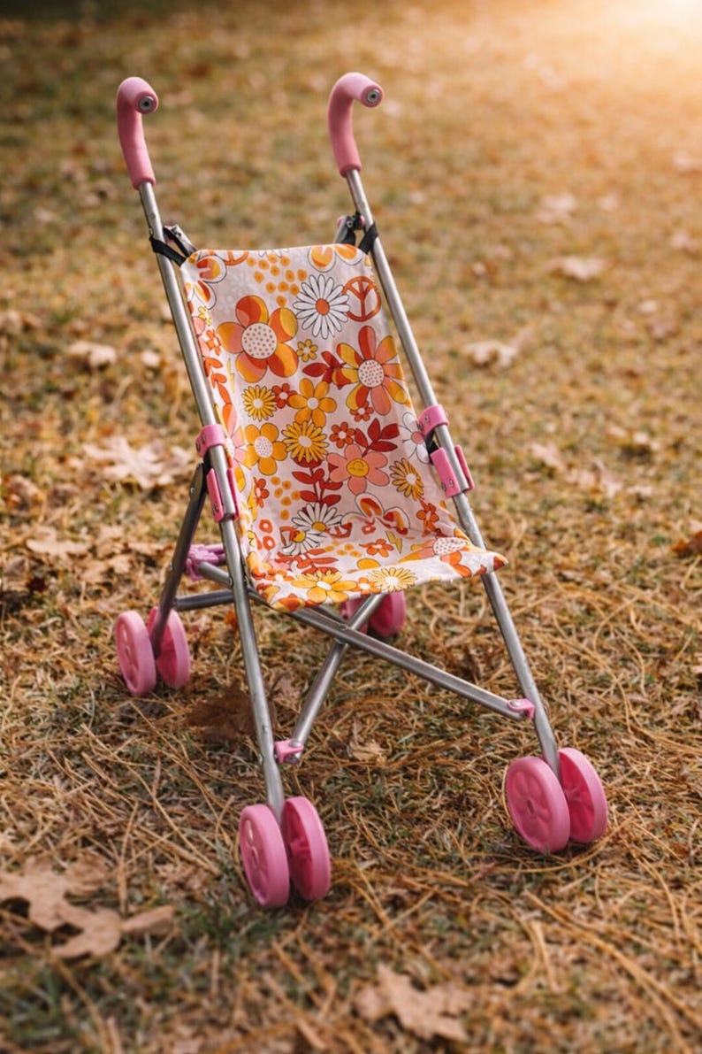 May include: A toy doll stroller featuring a metal frame with pink plastic handles and wheels. The seat fabric displays a repeating pattern of orange, yellow, and white flowers, along with peace signs, on a light background with a simple folding design.