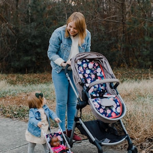 May include: A gray stroller featuring a seat liner with a dark blue floral pattern of pink and white flowers. A smaller toy stroller with a matching floral seat liner holds a doll with dark skin, wearing a pink shirt and blue denim shorts.
