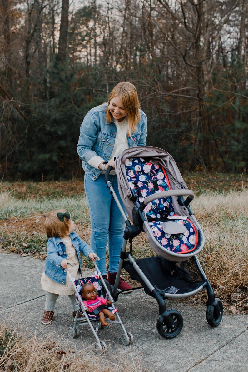 May include: A gray stroller featuring a seat liner with a dark blue floral pattern of pink and white flowers. A smaller toy stroller with a matching floral seat liner holds a doll with dark skin, wearing a pink shirt and blue denim shorts.