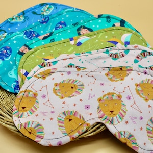 May include: Five colorful sleep masks with zodiac signs and constellations printed on them. The masks are made of soft fabric and have a black trim. The masks are arranged in a stack, with the top mask showing a pink background with a lion and stars.