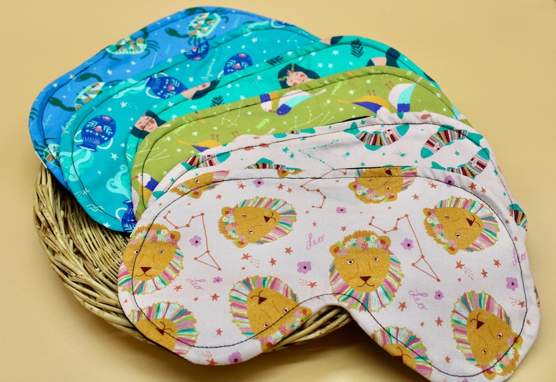 May include: Five colorful sleep masks with zodiac signs and constellations printed on them. The masks are made of soft fabric and have a black trim. The masks are arranged in a stack, with the top mask showing a pink background with a lion and stars.