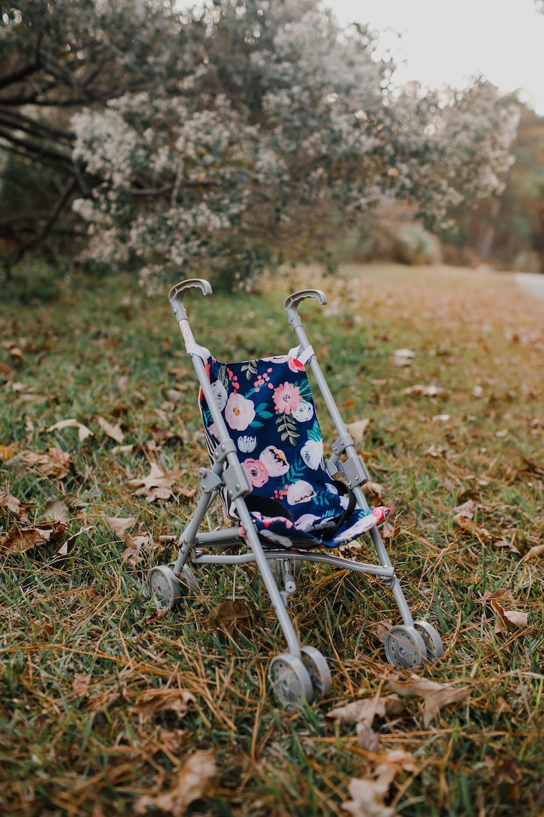 May include: A toy doll stroller featuring a navy blue fabric seat with a colorful floral pattern of pink, white, and light blue flowers and green leaves. The stroller has a light gray metal frame, two curved handles, and four sets of small wheels.