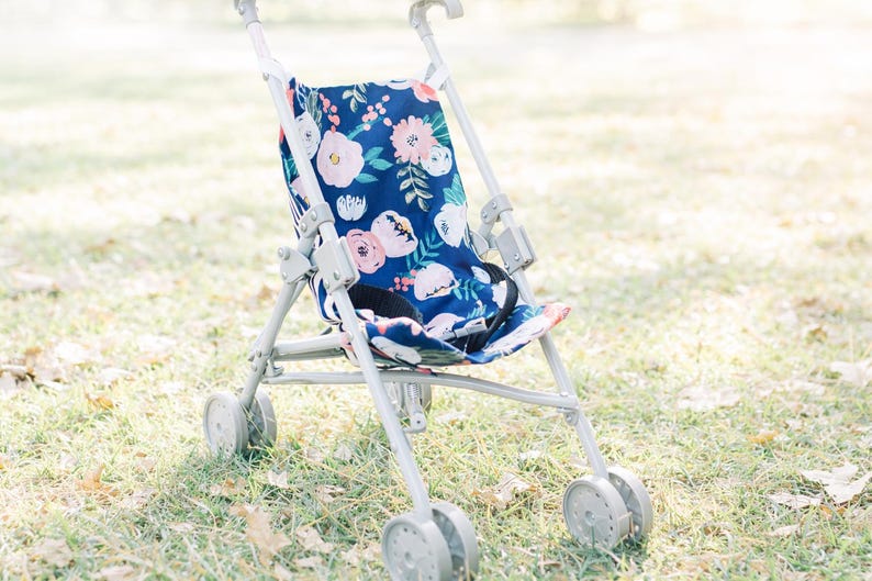 May include: A toy stroller featuring a navy blue fabric seat with a floral pattern of pink, white, and peach flowers and green leaves. The stroller has a light gray metal frame, four sets of double wheels, and a black safety strap across the seat.