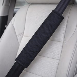 May include: A black seat belt shoulder pad is shown in a car. The pad is made of a soft, plush material and is designed to provide comfort and protection. The car seat is white leather.