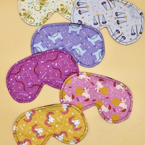 May include: Five different zodiac-themed sleep masks. Each mask is made of soft fabric and features a different zodiac sign, including Aries, Taurus, Gemini, Cancer, and Leo. The masks are all in a light color scheme with gold accents.