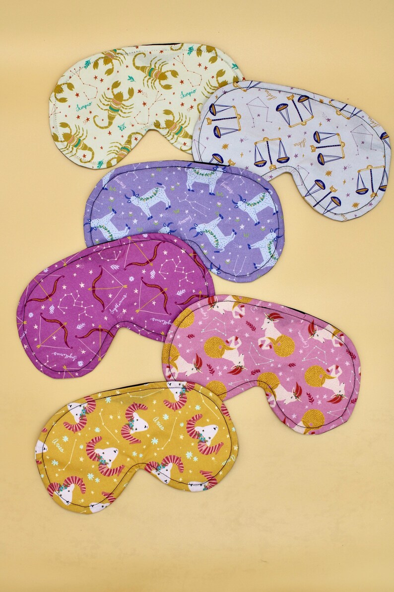 May include: Five different zodiac-themed sleep masks. Each mask is made of soft fabric and features a different zodiac sign, including Aries, Taurus, Gemini, Cancer, and Leo. The masks are all in a light color scheme with gold accents.