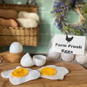 May include: A pack of white eggs labelled "Farm Fresh Eggs" with a black silhouette of a hen. Two white plastic eggs are open, and two felt eggs with yellow yolks are on a wooden chopping board.
