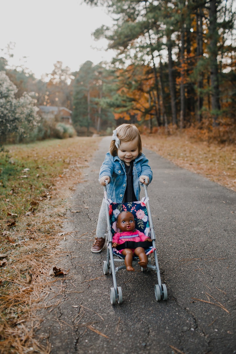 May include: A toy stroller featuring a dark blue fabric with a colorful floral pattern, containing a dark-skinned doll wearing a pink shirt with blue stripes. The stroller has a grey metal frame with four small wheels and a black safety strap.