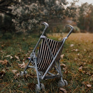 May include: A toy stroller featuring a grey plastic frame and a fabric seat with a black and white vertical striped pattern. The stroller has two curved handles at the top, a folding mechanism, and four sets of small wheels at the base.