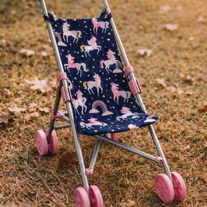 May include: A toy stroller with a silver metal frame and pink plastic handles and wheels. The seat fabric is dark blue and features a repeating pattern of pink and white unicorns, colorful rainbows, and yellow stars across the entire surface.