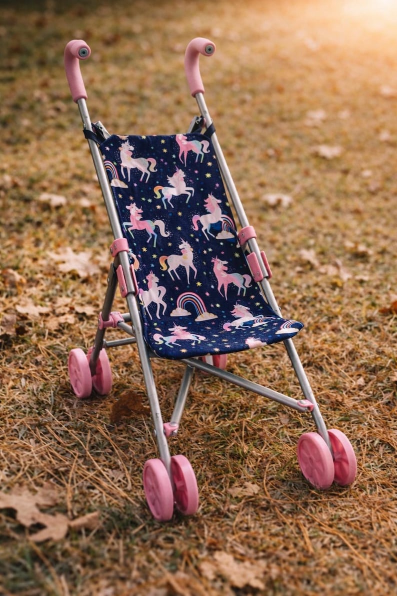 May include: A toy stroller with a silver metal frame and pink plastic handles and wheels. The seat fabric is dark blue and features a repeating pattern of pink and white unicorns, colorful rainbows, and yellow stars across the entire surface.