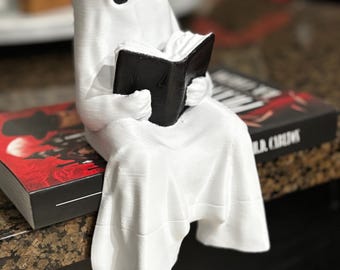 May include: A white ghost figurine with black eyes is seated, holding a black book. The ghost is draped in a sheet-like form, giving it a classic, spectral appearance. The figurine is placed on a book with red and black cover.