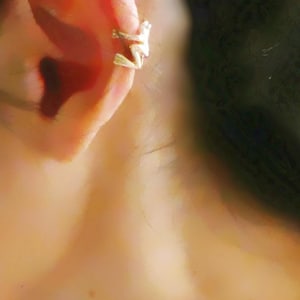 May include: A close-up of a person's ear with a gold ear cuff. The ear cuff is shaped like a curved line with a small, pointed end.