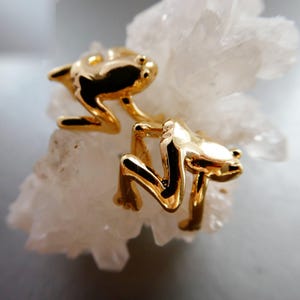 May include: A pair of gold-colored frog-shaped earrings. The earrings are detailed with visible eyes and legs, and are positioned on a white, crystalline surface. The frogs appear to be in a jumping pose.