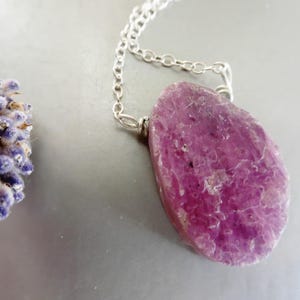 May include: A silver chain necklace featuring a large, irregularly shaped, polished pink gemstone pendant. The pendant has a textured surface and is a vibrant shade of pink. A sprig of lavender is in the background.