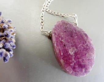 May include: A silver chain necklace featuring a large, irregularly shaped, polished pink gemstone pendant. The pendant has a textured surface and is a vibrant shade of pink. A sprig of lavender is in the background.
