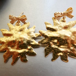 May include: A pair of gold-toned earrings featuring a sunburst design with a textured, hammered surface. Each earring is topped with a small, detailed bow accent. The earrings have a post-back closure and a bright, reflective metallic finish.