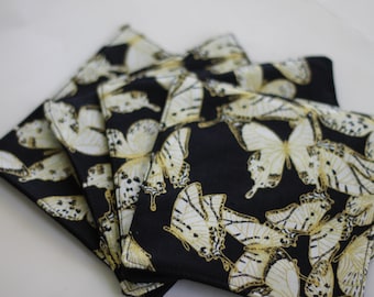 Puede incluir: Juego de cuatro posavasos de tela negra con un patrón de mariposa dorada y blanca.