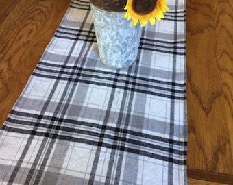 May include: A white and gray plaid table runner with black accents. The runner is on a wooden table with a vase of sunflowers in the center.