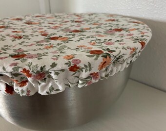 Puede incluir: Una cubierta de cuenco blanca con un estampado floral de flores rojas, naranjas y rosas. La cubierta tiene un borde con volantes y está colocada sobre un cuenco de acero inoxidable. El cuenco está sobre una estantería blanca.