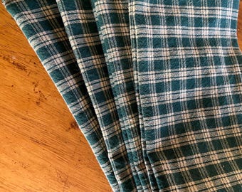 Puede incluir: Primer plano de varias servilletas de tela a cuadros verdes y blancos dobladas. Las servilletas están hechas de un material suave y están dispuestas sobre una superficie de madera. El patrón de cuadros es un diseño clásico, adecuado para diversas ocasiones.
