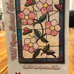 May include: A quilt pattern booklet titled Wild Roses Stained Glass by Bear Paw Productions. The cover features a graphic of pink flowers with yellow centers and green leaves, designed to look like stained glass, with the text Quilt As You Go Stained Glass.