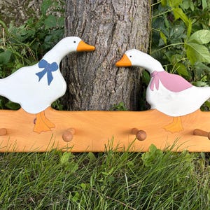 May include: A wooden wall hook rack featuring two painted white geese with orange beaks and feet. The left goose wears a blue bow, and the right goose wears a pink bow. The rack has four wooden pegs extending from the front of the light brown board.