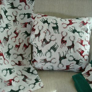 May include: A white fabric set featuring a repeating pattern of deer with red and green plaid accents. The set includes a pillow, a folded blanket, and two green towels with matching deer pattern trim.