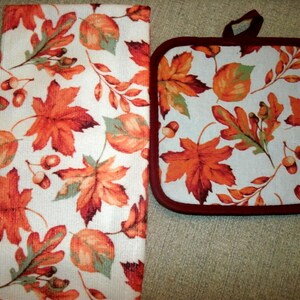 May include: A white kitchen towel and pot holder set with a fall leaf pattern. The design features orange, red, and green leaves, along with acorns, on a white background. The pot holder has a dark red border and a hanging loop.