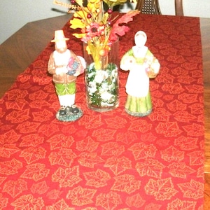 May include: A wooden dining table with a red leaf-patterned table runner. Two ceramic figurines and a vase of flowers are centered on the runner. The figurines depict a man and a woman in traditional attire.
