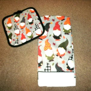 May include: A gray pot holder and matching towel set featuring a gnome pattern. The gnomes have red, orange, green, and black hats with white beards. The towel has a white border and the pot holder has a black border.