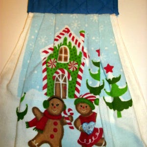 May include: A white kitchen towel with a gingerbread house, two gingerbread people, and a candy cane design. The towel has a blue quilted potholder attached to the top.