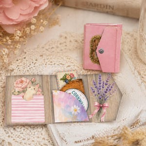 May include: A pink miniature wallet with a floral design and a button closure sits near a wooden display with decorative cards. The display features a cat illustration, flowers, and a lavender bouquet.