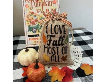 May include: A burlap bag with the text "I LOVE Fall MOST OF ALL" in black script, decorated with fall leaves. The bag is tied with a plaid ribbon and sits on a white doily. Decorative pumpkins and a sign that says "Pumpkin Farm" are in the background.