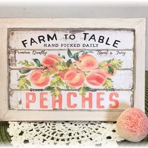 Puede incluir: Un letrero de madera enmarcado con las palabras "Farm to Table" y "Peaches" en un estilo vintage. El letrero presenta una ilustración de acuarela de melocotones y hojas. Un melocotón se coloca frente al letrero.