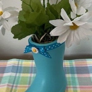 May include: A turquoise boot-shaped vase filled with white daisy flowers and green leaves. A blue polka-dot bow with a daisy accent adorns the vase. The vase is sitting on a plaid surface.