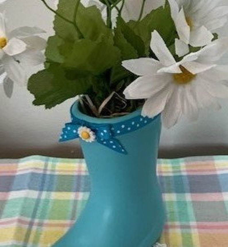 May include: A turquoise boot-shaped vase filled with white daisy flowers and green leaves. A blue polka-dot bow with a daisy accent adorns the vase. The vase is sitting on a plaid surface.