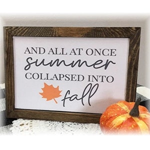 May include: A rectangular wooden sign with the text "AND ALL AT ONCE summer COLLAPSED INTO fall" in black script. An orange leaf graphic is below the word "summer". A small orange pumpkin sits to the right of the sign.