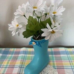 May include: A light blue boot-shaped vase filled with white daisy flowers. The vase has a blue polka dot ribbon with a yellow flower accent. The arrangement sits on a colorful plaid tablecloth.