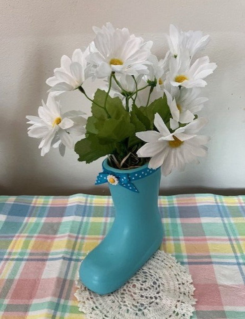 May include: A light blue boot-shaped vase filled with white daisy flowers. The vase has a blue polka dot ribbon with a yellow flower accent. The arrangement sits on a colorful plaid tablecloth.
