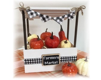 May include: A white wooden farmers market stand with a black and white checkered banner and trim. The stand is filled with various orange and yellow pumpkins. A small chalkboard sign reads "Farmers Market".