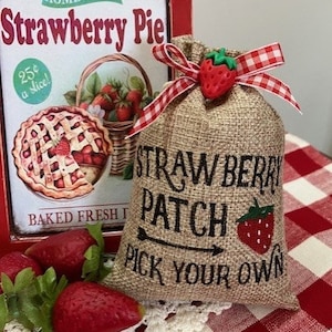 Pode incluir: Um saco de juta com uma fita xadrez vermelha e branca e um amuleto de morango vermelho. O saco é decorado com o texto "Straw Berry Patch" e uma seta apontando para a direita com o texto "Pick Your Own".