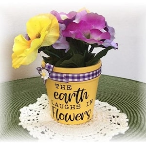 May include: Yellow ceramic flower pot with purple and white gingham ribbon. The pot has a black painted design that says "The earth laughs in flowers".