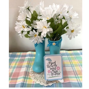 May include: A spring-themed arrangement featuring white daisy flowers in light blue rain boots. The boots are decorated with blue ribbon and a daisy accent. A small sign reads "April Showers bring May flowers."