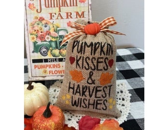 May include: A burlap bag with the text "Pumpkin Kisses & Harvest Wishes" in black script, decorated with orange leaf and heart accents. The bag is topped with a small orange pumpkin and a plaid ribbon. A pumpkin farm sign is in the background.