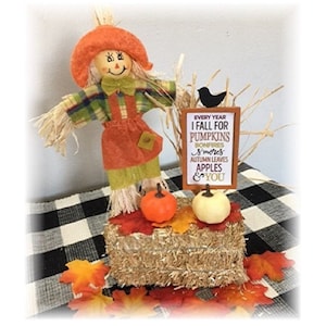 Könnte beinhalten: Herbstliche Dekoration mit einer Strohpuppe mit orangefarbenem Hut und Latzhose, die auf einem Heuballen steht. Die Dekoration umfasst Kürbisse, Herbstblätter und ein Schild mit der Aufschrift "I fall for pumpkins".